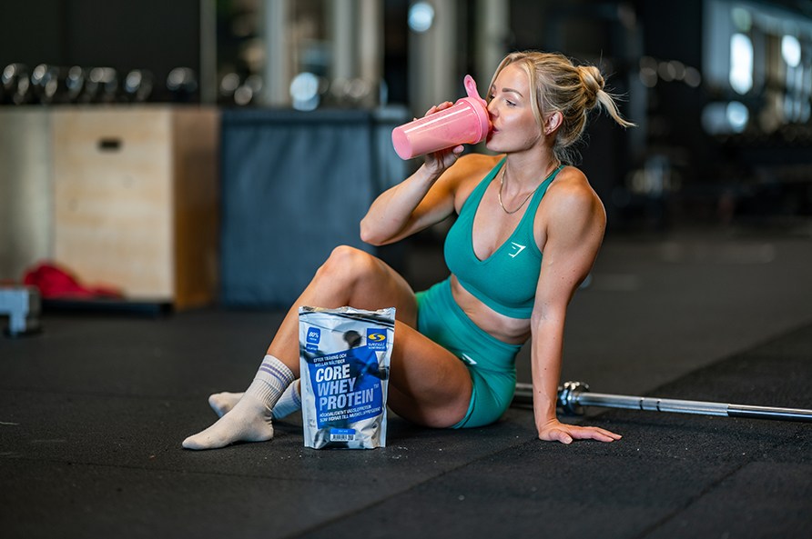 Kvinna sitter p golvet i ett gym och dricker Core Whey Protein ur en shaker.